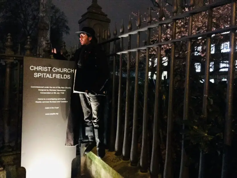 Nighttime Jack the Ripper Tour London. Guide standing near the lit Christ Church Spitalfields sign, in front of a black iron fence