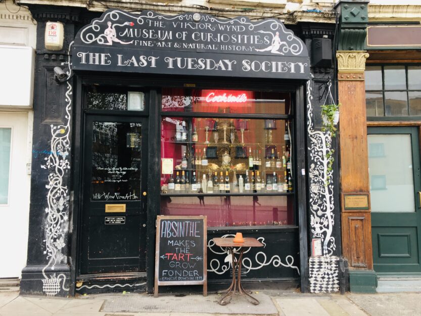 Front window  full of glass alcohol bottles of Viktor Wynd Museum of Curiosities in London, it has black woodwork with white writing that says 'the Viktor Wynd Museum of Curiosities. Fine Art & Natural History. The Last Tuesday Society'