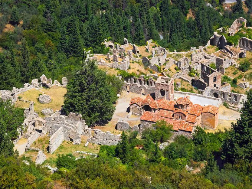 View from above of the ancient town of Mystras. Mainly grey stone ruined wallks and fortifications and a church wtith a red tiled roof. Surrounded by green evergreen trees.