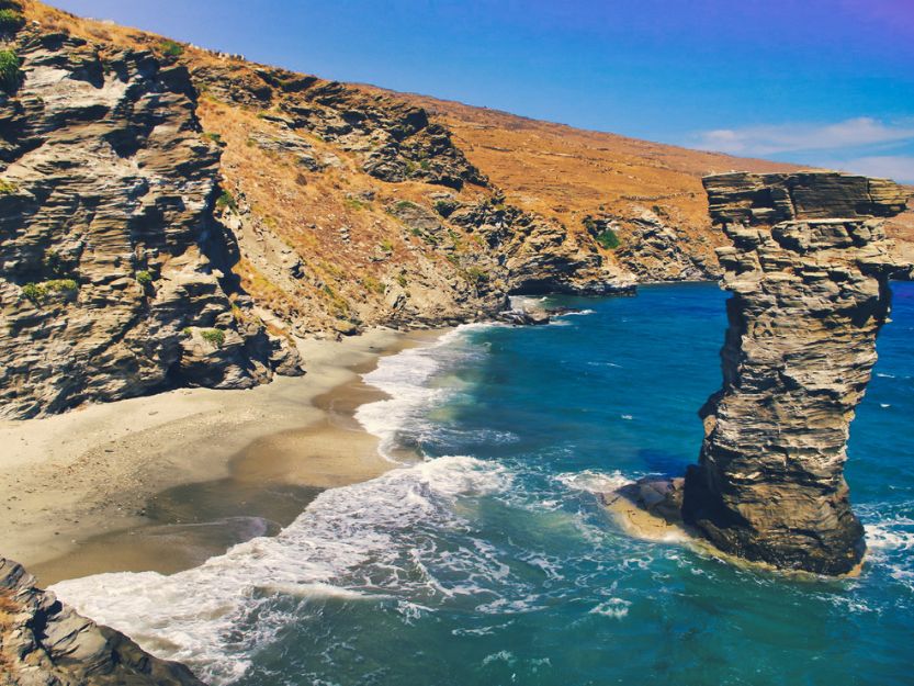 Andros a Cyclades Island in Greece. Image of beach with  yellow sand beach red rocks to the left and blue sea and rock column to the right