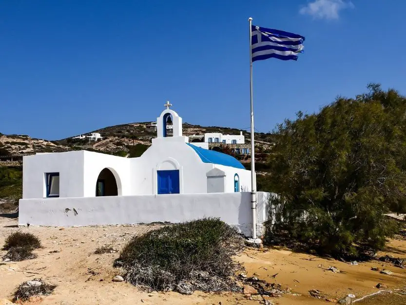 Antiparos, a Cyclades Island in Greece. Blue and white church with the Greek flag to the right and green hills and vegetation.