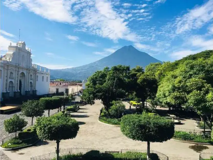 Central Park in Antigua, Guatemala, wirh shaped trees and volcano in the background under a blue sky with white clouds.