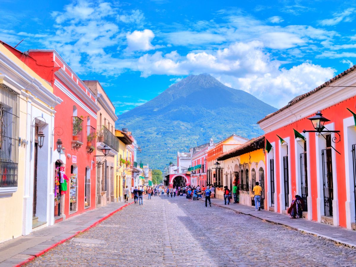 Things to do in Antigua, Guatemala. A cobbled street, lined both sides with bright colourful houses and a volcano and cloudy blue sky in the background.