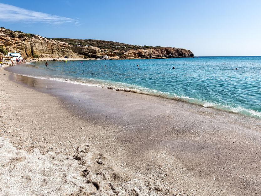 Firiplaka Beach on Milos in Greece. Fie sand beach on the left curving gently towards rocks at the end of the bay with blue sea to the right.