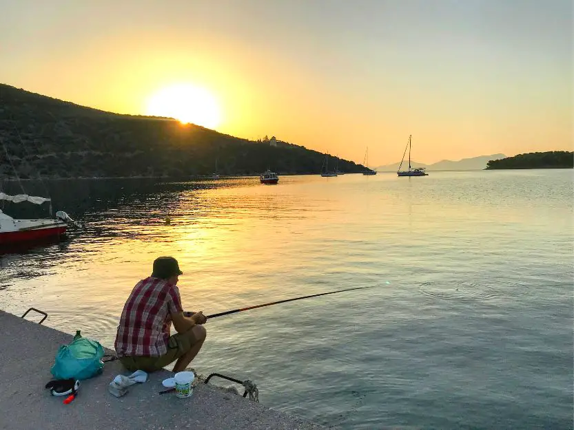 Andy fishing at sunrise, sitting on the port on Limani side of Ermioni. He is wearing a red and white checked shirt.