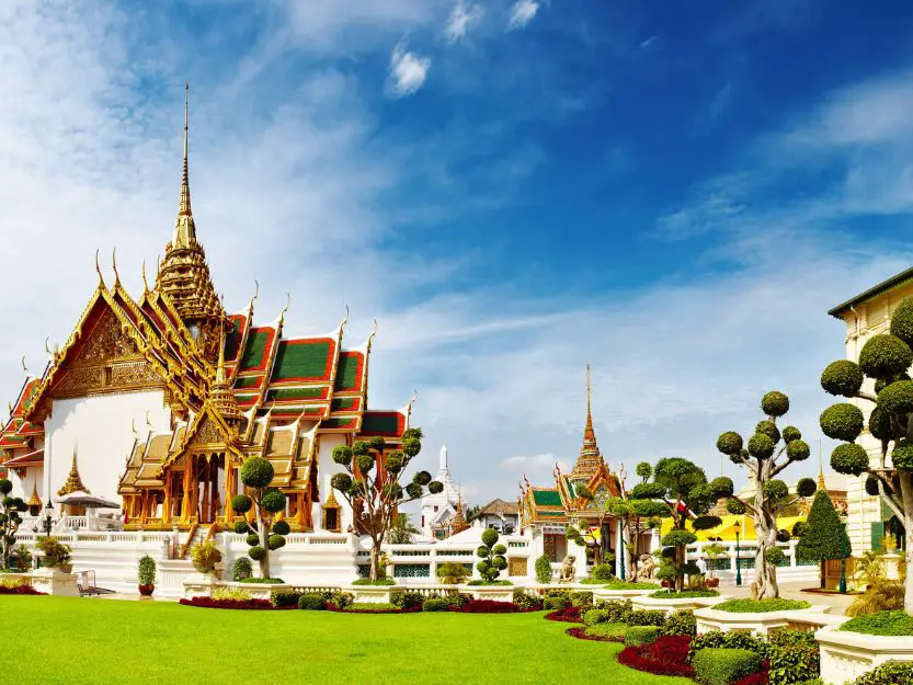 Grand Palace in Bangkok, Thailand. White walls, green, gold and red roof, with green grass in foreground and blue sky in the background.