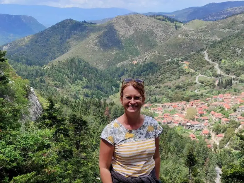 Tanya standing with the view of Tsintzina village, the valley and mountains behind her on a short hike to Agios Ioannis in the Paronas Mountains, Laconia. She's wearing a white and yellow striped t-shirt and has her red hair tied back.