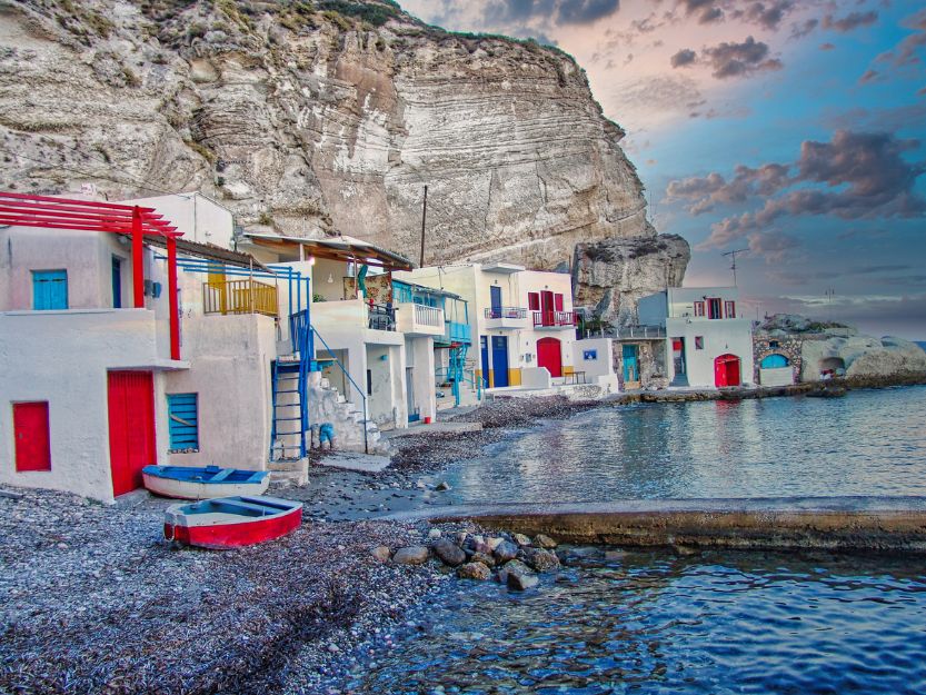 White and colourful Syrmatas (old fishing houses) on the pebbly Kilma Beach fishing village. Cliffs towering above on the left and calm sea on the right.