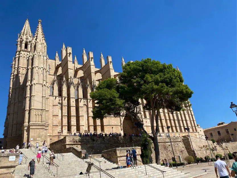 Palma de Mallorca Cathedral, with blue sky in background and green tree and people walking in foreground