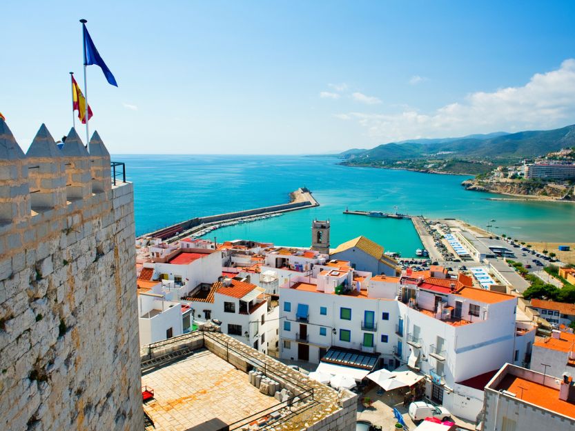 View of the Peniscola Port in Valencia, Spain. Castle turrets in the foreground, overlooking white houses with terracotta roofs, with blue sea and mountains in the background.