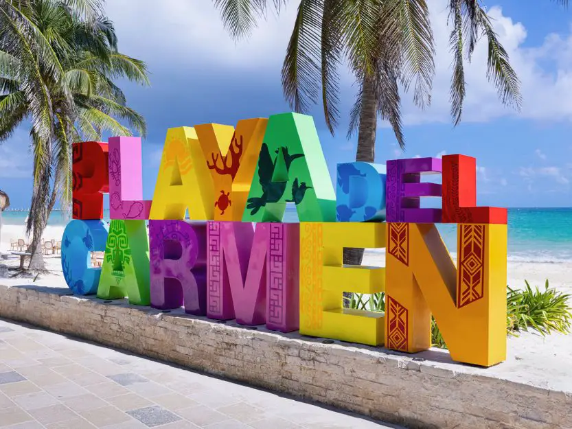 Colourful Playa del Carmen sign with two palm trees, white sand beach and blue sea behind it.