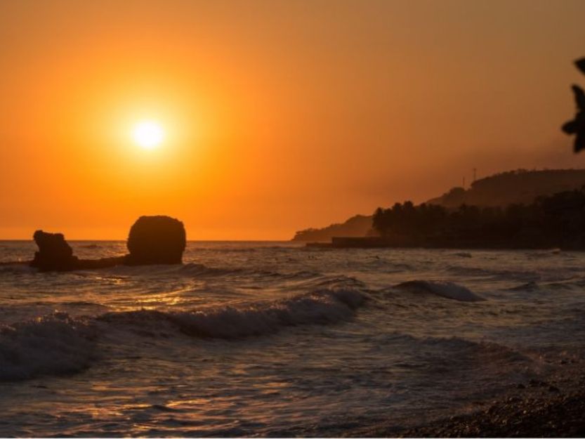 Sunset at Playa El Tunco, orange sunset behind pig rock. With hills in the background and waves crashing on the rocks in the foreground.