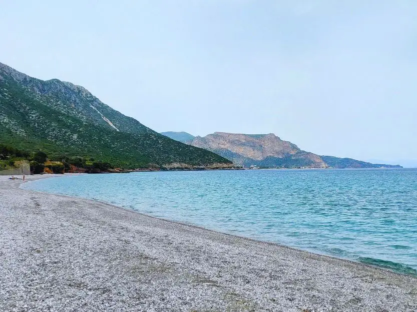 Curved pebbled Poulithra Beach with blue sea to the right and vegetation covered mountains to the right