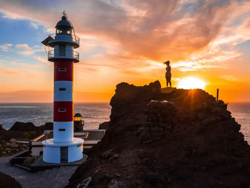 Punta de Teno Lighthouse. Image of red and white lighthouse at sunset with a woman standing on a rock.