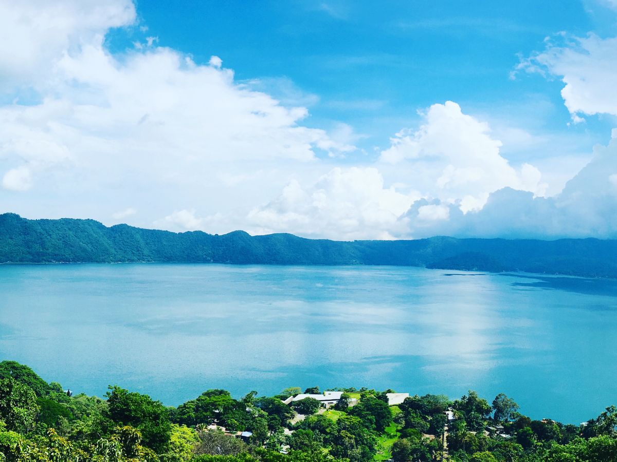 Reasons Not to Skip Lake Coatepeque, El Salvador. Image of blue lake in a crater surrounded by green trees, beneath a blue sky with white clouds