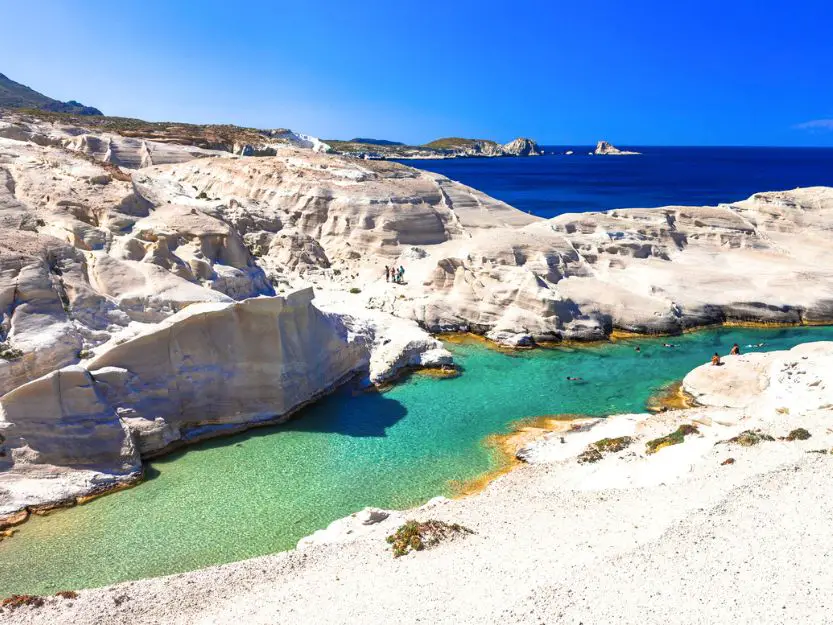 Sarakiniko Beach on Milos in Greece. Lunar like light beige rocks, surrounding a clear turquoise blue swimming area with darker blue sea in the background and blue sky above.