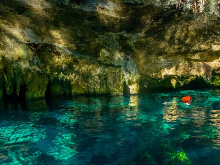 Person wearing a red life jacket, snorkelling in the clear blue water of Gran Cenote in Tulum in mexico. The sinkhole also has stalagmites and stalactites.