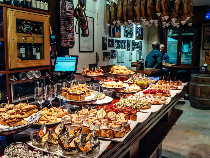 Lots of tapas dishes in a bar in San Sebastian, Spain. Hams are hanging from the ceiling and two men sitting in the background.