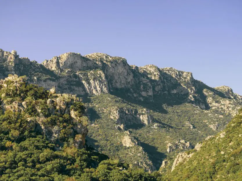 Taygetus mountains in Laconia. Light grey limestone rock mountain range, covered in patches of green trees and bushes. Light blue sky in the background.