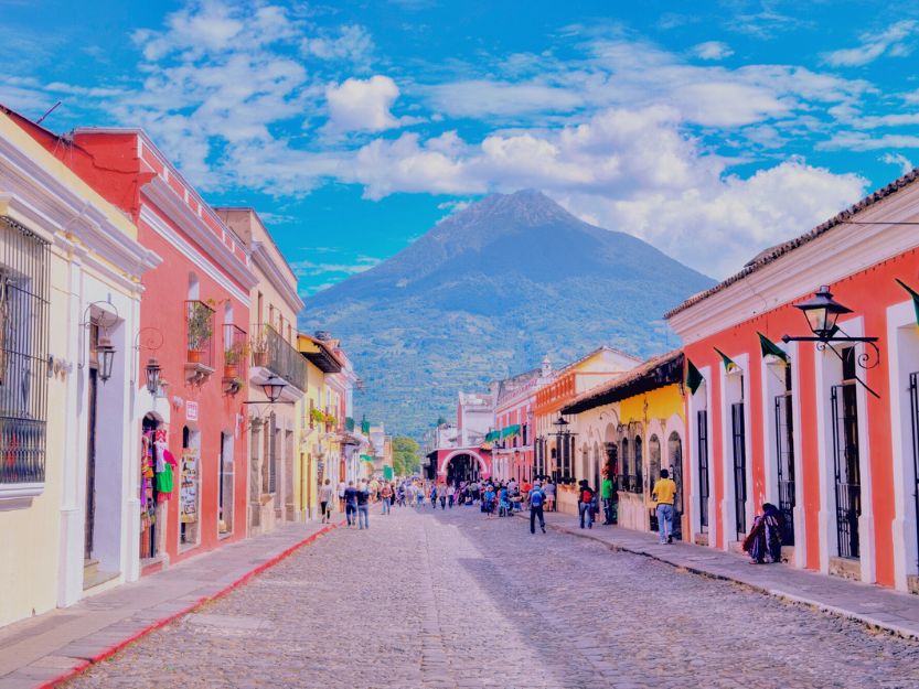 Things to Do in Antigua Guatemala. Cobbled street lined with colourful houses with volcano and blue sky with white clouds in the background.