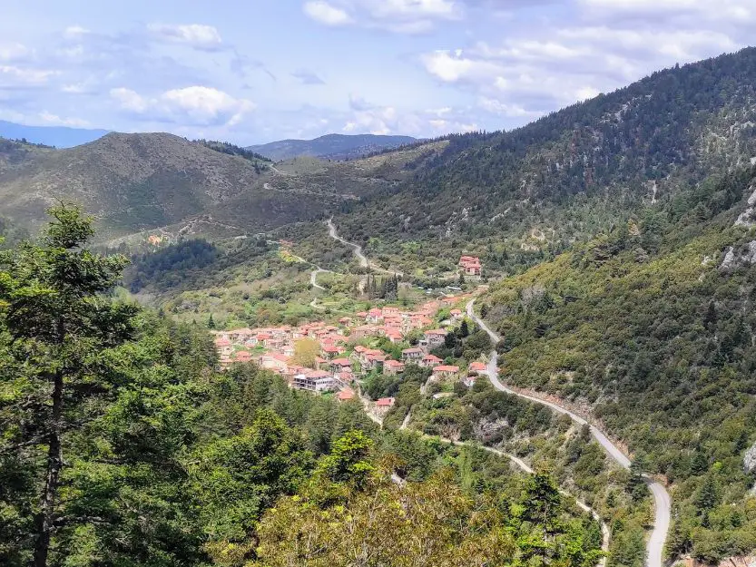 Things to do in Laconia, Greece. An aerial view of Tsintzina village nestled amongst green slopes covered in fir trees. Mountains and sky with clouds in the background.