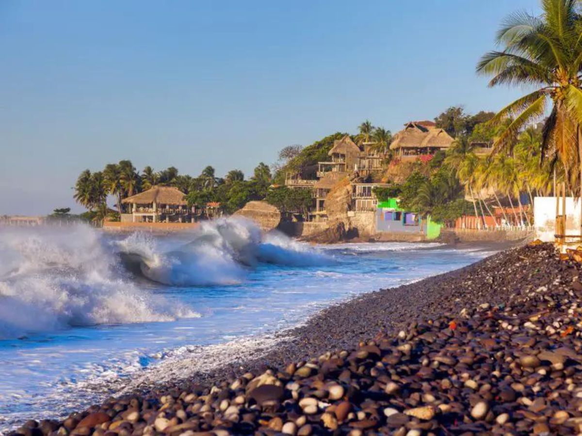Things to do in Playa El Tunco. Image of pepple beach with big surf waves crashing on the shore. To the right on the land are palm trees and palm leaf roof buildings.