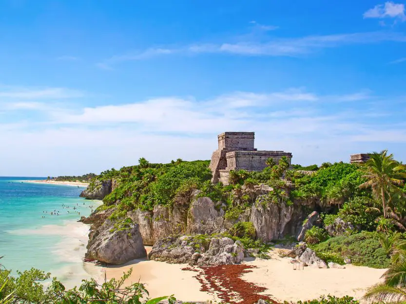 Things to do in Tulum in Mexico. Image of the clifftop ruins on the right overlooking a white sand beach and turquoise blue sea on the left. The sky is blue with white wispy clouds.