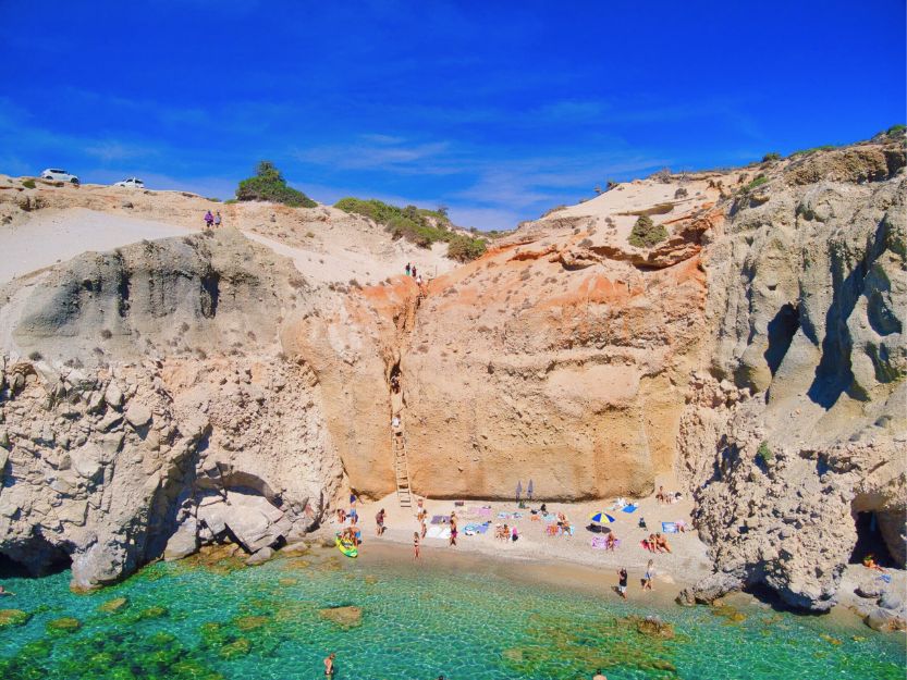 Tsigrado Beach on Milos Greece. Small sandy beach with bright blue sea surrounded by tall red stone cliffs.