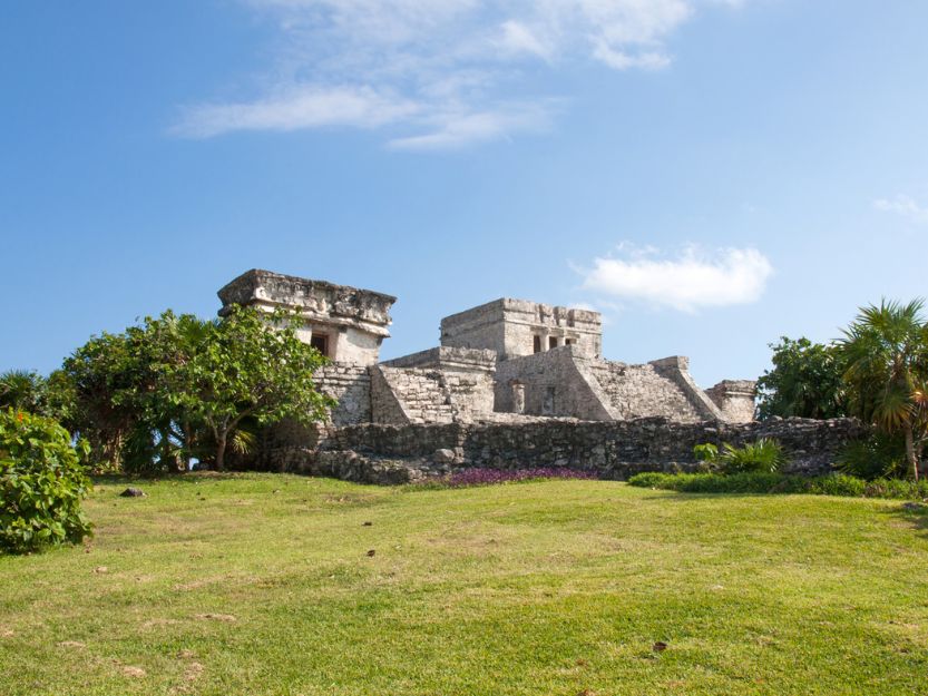 Tulums cliffside ruins. Grey stone mayan ruins, surrounded by green palm trees and grass in the foreground. Blue sky with white wispy clouds in the background.