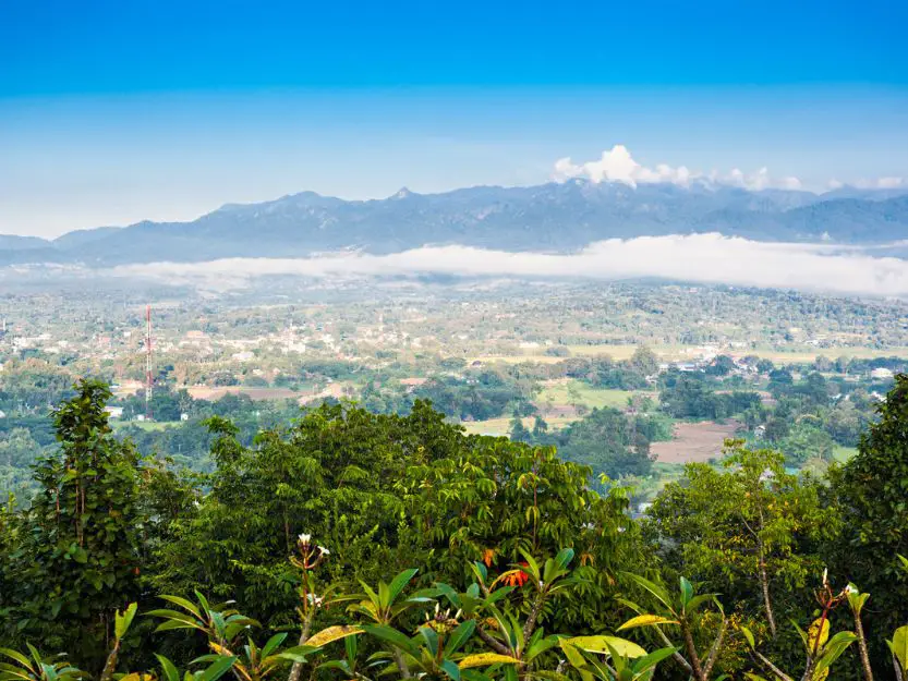 Aerial view of Pai in Thailand, with blue sky and cloud topped mountains in the background, then the village in front and vibrant green trees in the foreground.