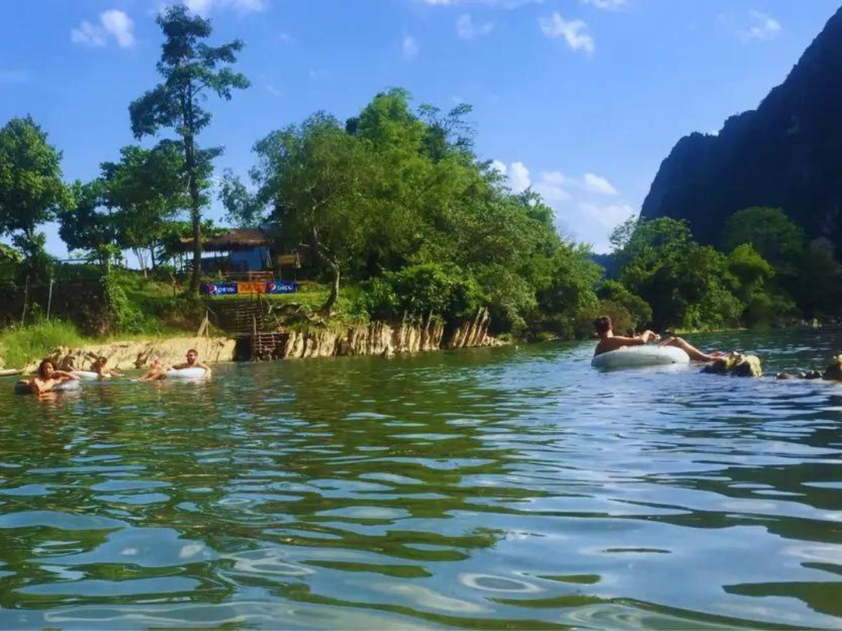 Vang Vieng tubing in Laos. Tourists floating in white rubber rings on the river which is lined with grass and green trees and a mountain in the background to the right.