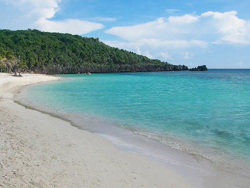 West Bay Beach on Roatan Island in Honduras. White sandy bay beach on left with clear turquoise sea on the right with green tree covered hill in background.