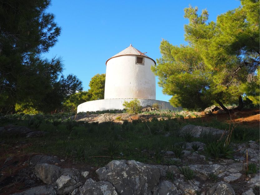 Old white windmill on the top of the Bisti in Ermioni. Surrounded by green pine trees with rocks in the foreground and blue sky above